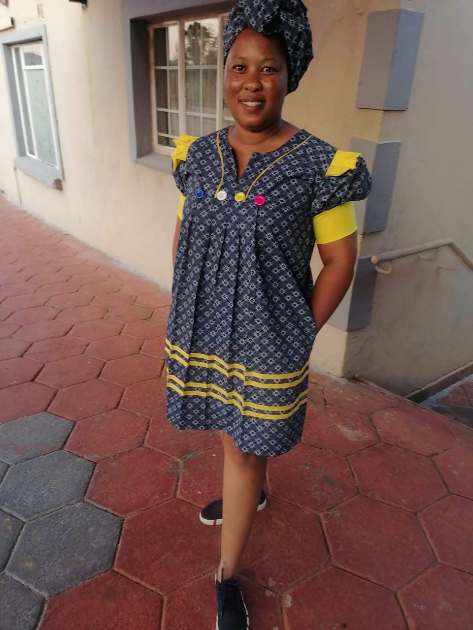 Woman wearing a navy shweshwe pleated shift dress with yellow trim, matching headwrap and coloured buttons