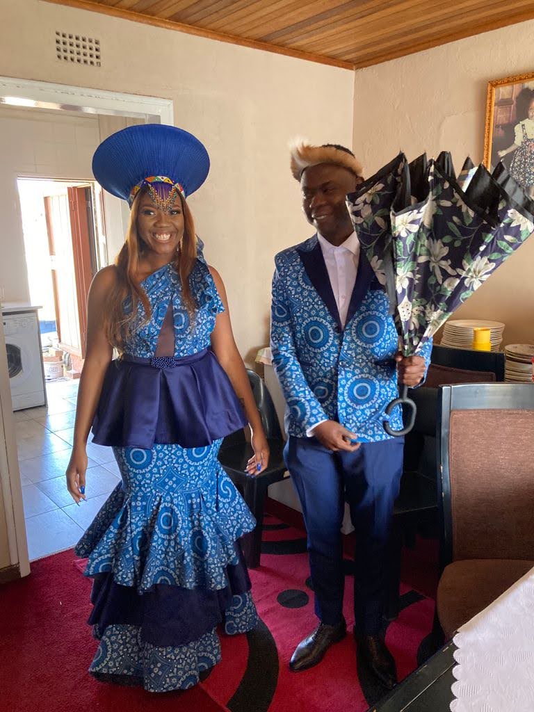 Bride in a tiered blue shweshwe mermaid skirt and satin peplum top beside her groom in a matching printed blazer