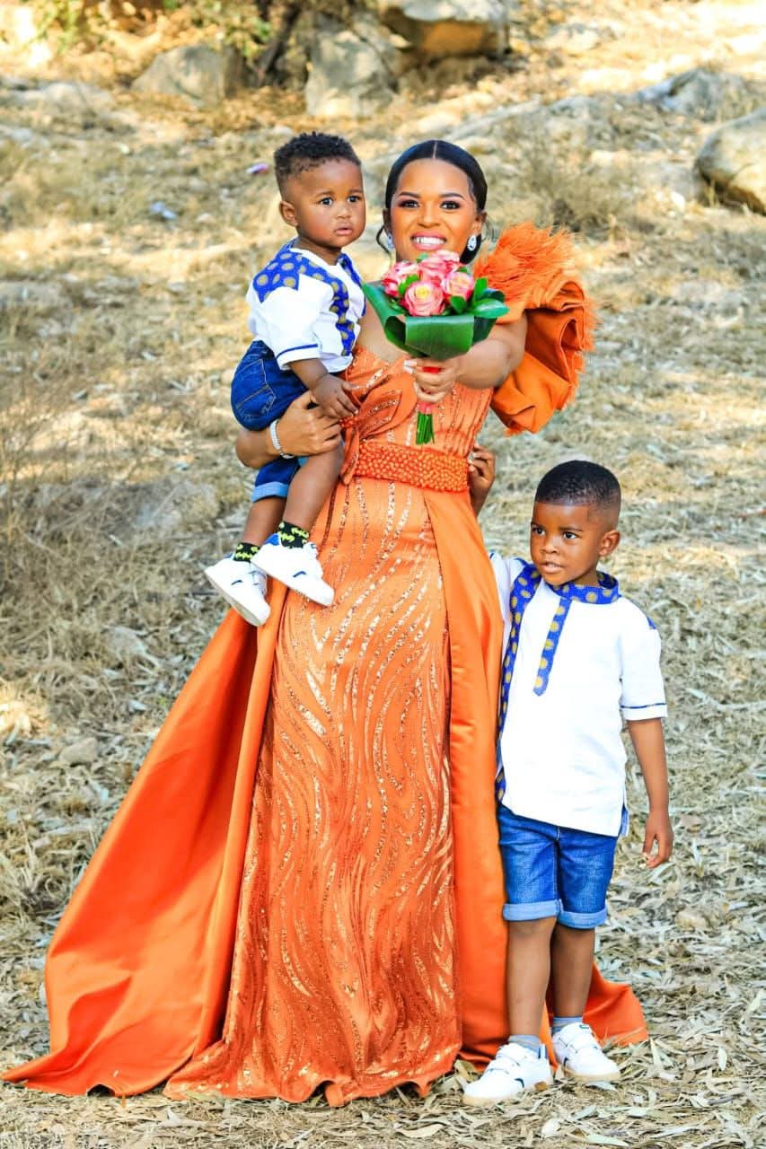 Bride in a burnt orange glitter bridal gown with beaded waist and satin train holding her two sons in matching shweshwe-trimmed shirts