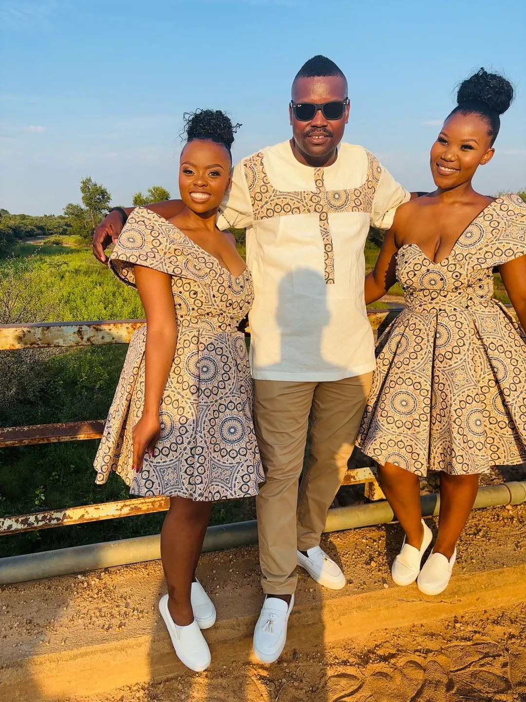Two women in matching flared shweshwe dresses standing with a man in a coordinating cream shweshwe-panelled tunic